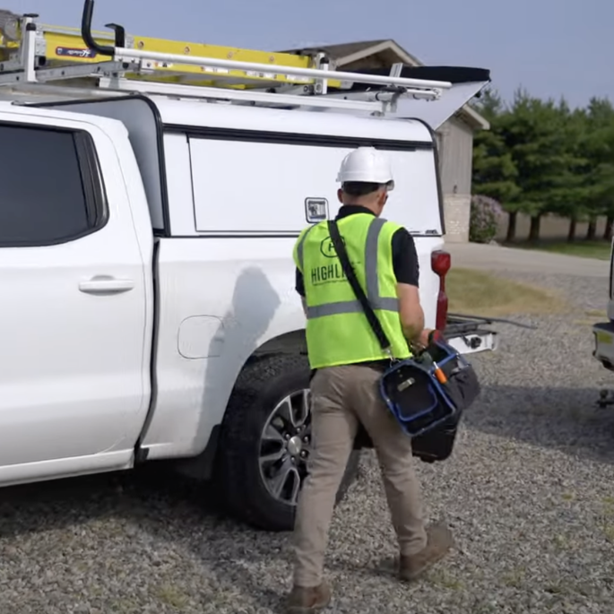 Man in a safety vest with a bag of equipment walking away from a truck.